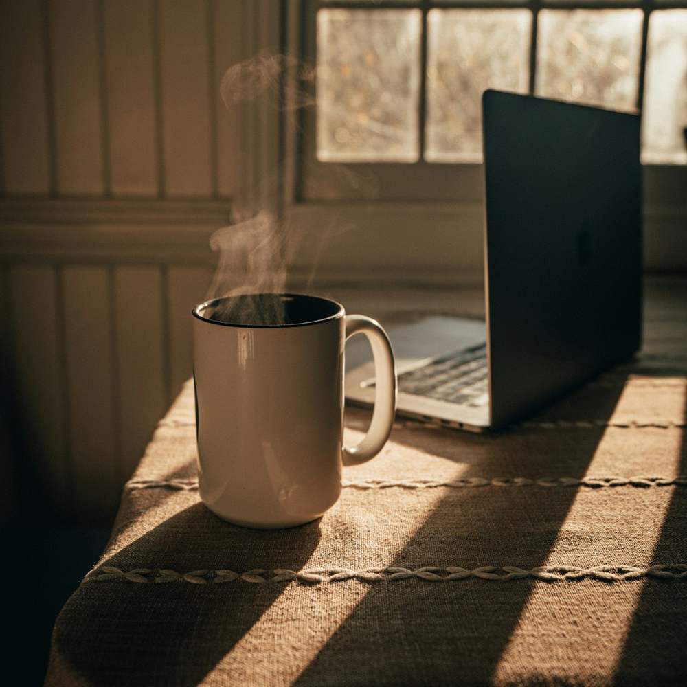 Šolja vruće kafe i laptop na stolu uz prozor, jutarnje sunce
